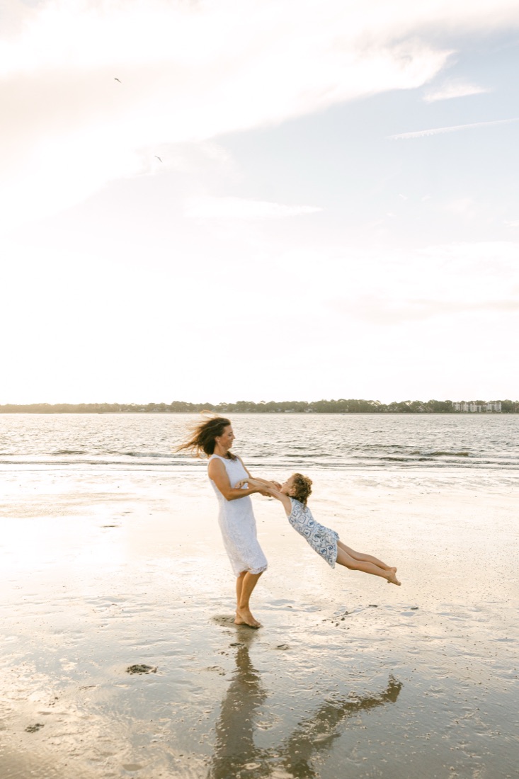Val swinging her daughter on a wide beach at sunset